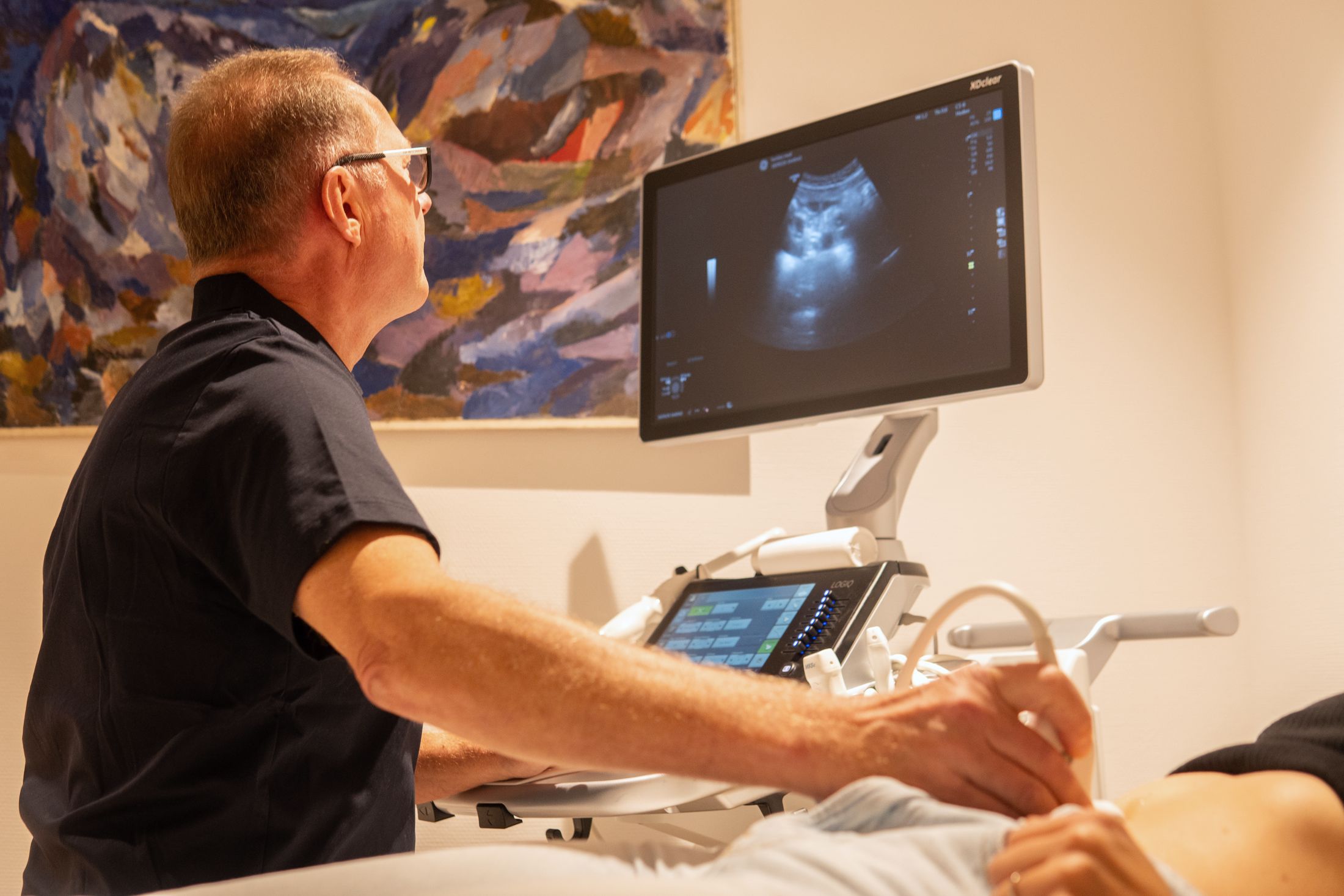 Dr. Balzer bei einer Ultraschalluntersuchung, im Hintergrund ein Gemälde